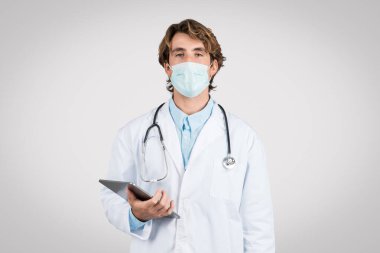 Cerrahi maske ve steteskop takan odaklanmış erkek doktor, dijital tablet kullanıyor, sağlık ve teknoloji kaynaşmasını işaret ediyor, gri arka plan