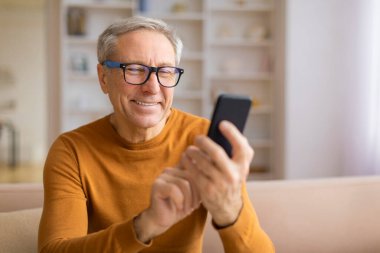 Gözlüklü, gülümseyen yaşlı bir adam akıllı telefonuna odaklanmış ev ortamında ilgi ve rahatlık taşıyor.