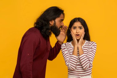 Bir erkek, bir kadının kulağına bir sır fısıldar. Kadının şok olmuş ifadesi, canlı sarı bir arkaplan karşısında büyüleyicidir.