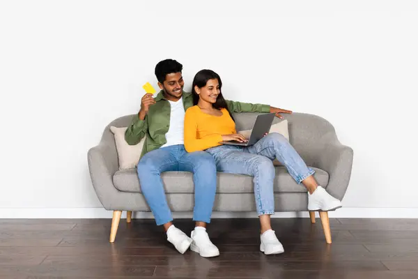 stock image Interested Indian couple engaged in online shopping, holding credit card and laptop while comfortably sitting on grey couch, in minimalistic space