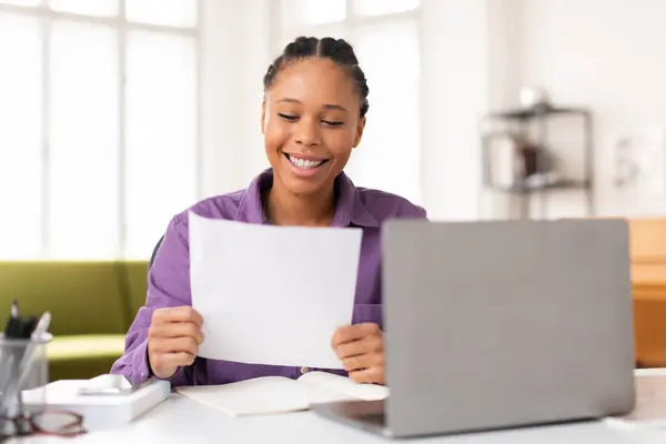 身穿紫色衬衫的快乐的黑人少女高兴地阅读文档 坐在办公桌前 打开笔记本电脑 四周都是学习材料 — 图库照片