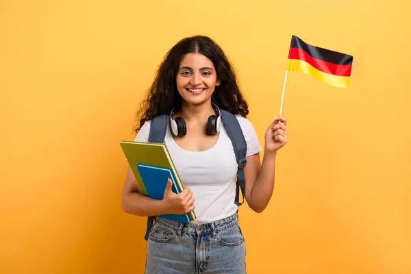ヘッドフォンを持つ幸せな女性学生は 外国語学習を示唆するドイツの旗とカラフルなノートブックを保持しています — ストック写真