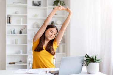 Mutlu Asyalı kadın kollarını kaldırarak esnetiyor. Ev bilgisayarında masa başında otururken işe ara veriyor. Başarılı bir iş gününden sonra gevşeyen neşeli Koreli kadın.
