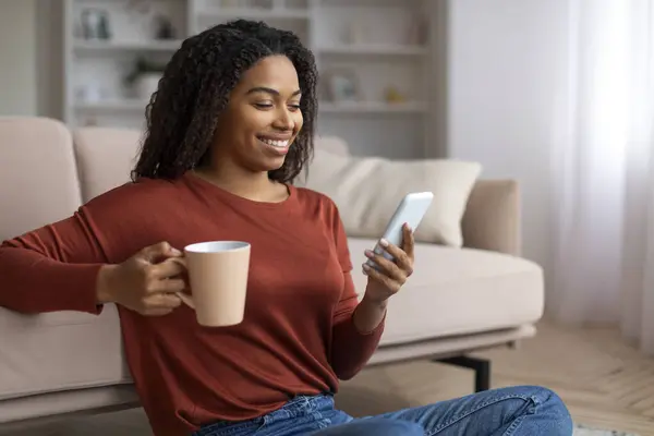 Domestic Leisure. Smiling Black Woman Relaxing On Floor With Smartphone And Coffee, Cheerful African American Female Browsing Social Networks On Mobile Phone And Enjoying Hot Drink, Copy Space