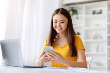 Gülümseyen Asyalı genç bir kadın ofisteki masasında otururken akıllı telefonunu karıştırıyor. Önünde dizüstü bilgisayarı açık, mutlu Koreli serbest çalışan kadın iletişim için cep telefonunu kullanıyor.