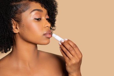 Siyahi bir kadın zarif bir şekilde dudak kremi sürüyor, sakin bir ifadeyle, tamamlayıcı bej arka plan, serbest alan üzerine kişisel bakım vurguluyor.