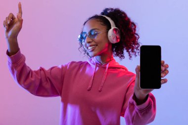 Kıvırcık saçlı neşeli bir kadın, güneş gözlüğü ve kulaklık takıyor, akıllı telefon boş ekranını gösterirken dans ediyor, parlak neon ışıklı arka planda,