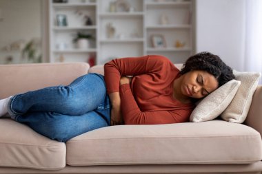 Genç siyahi kadın karnını evdeki rahatsızlıkla sıkıca tutuyor. Afro-Amerikan kadın adet sancısı ya da mide rahatsızlığı çekiyor. Oturma odasındaki kanepede yatıyor.