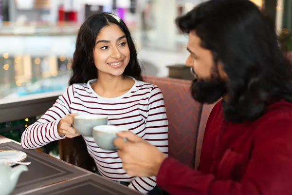 Beautiful millennial indian couple have romantic date at coffee shop, smiling eastern man and woman sitting next to each other, drinking tea, have conversation, cozy cafe interior