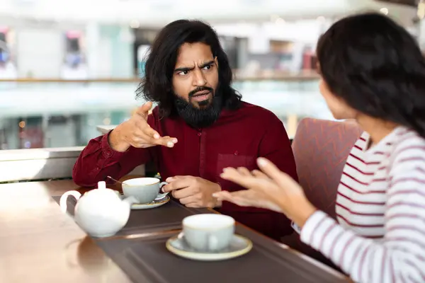 Angry emotional young indian couple have fight at cafe. Furious bearded eastern man talking rude and gesturing while have conversation with his girlfriend, cafe interior, copy space