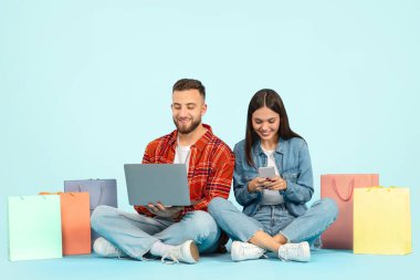 Modern alışveriş. Genç çift, alışveriş poşetleriyle bilgisayar ve akıllı telefonlardan online alışveriş yapıyor, mavi stüdyo arkaplanının karşısında oturuyor, tüketim ve ekommerizmi simgeliyor.