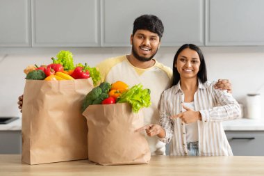 Market poşetlerini işaret eden mutlu Hintli çift, mutfak içlerinde duruyor, gülümseyen milenyum eşleri organik sebzeleri gösteriyor, market alışverişinden sonra poz veriyor, süpermarkete yiyecek teslimatı öneriliyor.