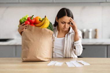Hayal kırıklığına uğramış, bin yıllık Hintli kadın. Elinde kağıt torba dolusu organik gıda ile market alışverişinden sonra faturaları kontrol ediyor. Yüksek fiyatlar, rahat mutfak iç dekorasyonu...