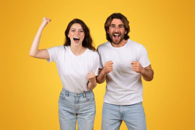 Güçlü Avrupalı genç çift, yumruklarını kaldırarak zaferi kutluyor, sevinç ve heyecan saçıyor, beyaz t-shirtler ve kot pantolon giyiyorlar, canlı sarı bir arka plan, stüdyo