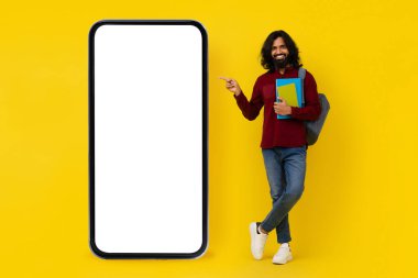 Sırt çantası taşıyan uzun saçlı ve sakallı Hintli adam ve beyaz ekranlı telefonu işaret eden kitap öğrencisi, güzel bir eğitim mobil uygulaması, sarı stüdyo arka planı, kopyalama alanı gösteriyor.