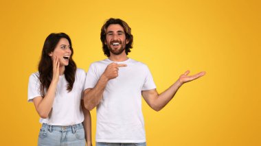 Neşeli Avrupalı çift etkileşime giriyor ve gülüyor, adam kadını işaret ediyor, sanki bir espri paylaşıyormuş gibi, ikisi de beyaz tişört giyiyor, sarı bir arka plana karşı neşeli bir anın tadını çıkarıyor.