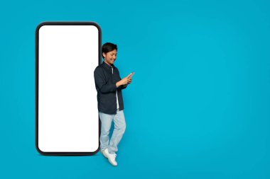 Mutlu Asyalı adam ayakta duruyor ve yanında kocaman boş bir akıllı telefon ekran modeliyle parlak bir turkuaz arka planda, bağlantı ve tasarım alanını gösteren bir telefon kullanıyor.