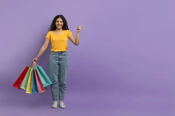 ショッピングバッグを持っている陽気な女性と紫色の背景に笑顔で上向きを指す — ストック写真