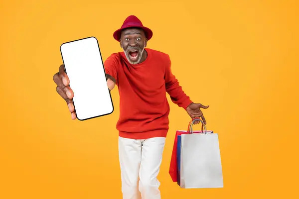 一名身穿红色毛衣 头戴红色帽子 面带微笑的年长黑人男子 拿着一个空白的智能手机屏幕 拿着五颜六色的购物袋 背景是橙色的工作室 — 图库照片