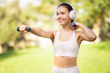 Odaklanmış, zinde, gülümseyen, spor kıyafetli, beyaz kulaklıklarıyla müzik dinlerken dambılları kaldıran beyaz tenli bir kadın.