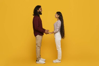 Bir erkek ve bir kadın el ele tutuşup, canlı sarı bir arka plana karşı durarak, bir bağ ve beraberlik hissini tasvir ederler.