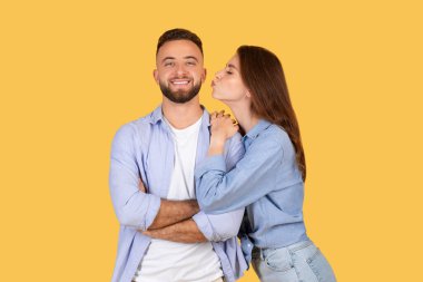 Gülümseyen bir adamı yanağından öpen bir kadın sarı bir arka planda hem rahat giyinmiş hem de mutlu görünüyor.