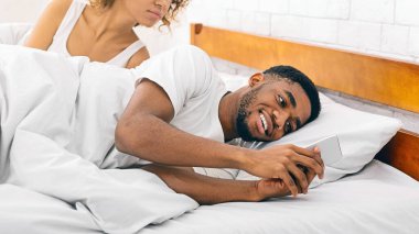 Dürüst bir güvensizlik anı, Afrikalı Amerikalı bir adamı yatakta habersiz ortağının yanında gizlice telefonunu kontrol ederken tasvir etmek.