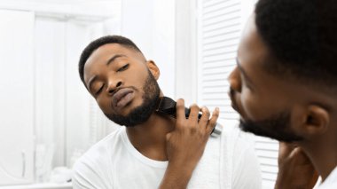 Odaklanmış Afrikalı Amerikalı adam sakal bırakıyor. İyi ışıklandırılmış, modern bir banyo ortamında budayıcı kullanıyor.