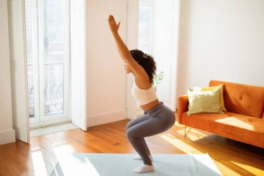 Kapalı mekanda kollarını kaldırarak çömelen genç bir bayan, ev içi antrenman yapıyor, oturma odasında aerobik ve pilates egzersizleri yaparak kaslarını güçlendiriyor.