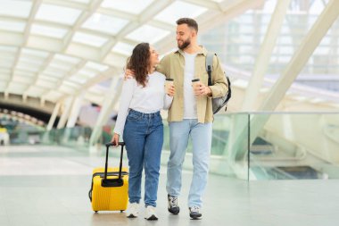 Çağdaş bir havaalanı ortamında gülümseyen ve birlikte yürüyen genç çift, seyahat ve mutluluk hissini aktarıyor.
