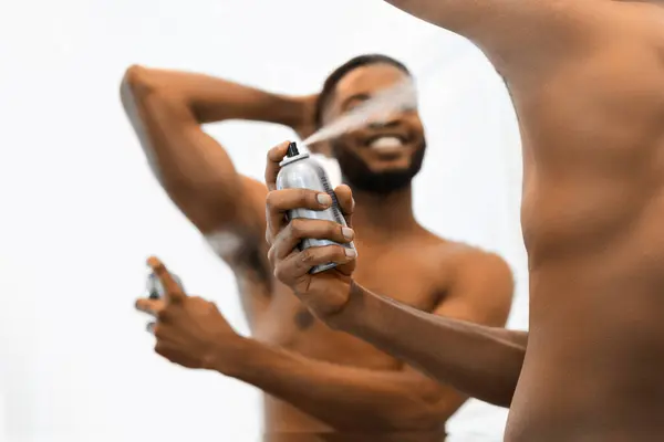 白い背景にある筋肉のないシャツのないアフリカ系アメリカ人男性は 消臭剤ボトルを保持し 腕の下に噴霧し 個人的な衛生状態を示しています — ストック写真