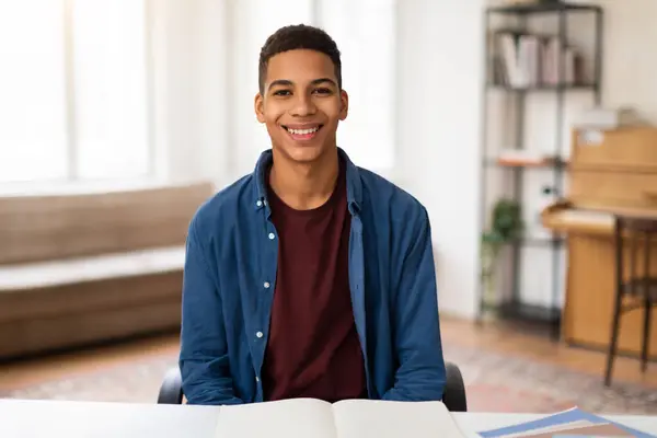 身穿蓝色衬衫 面带微笑的黑人男生坐在课桌前 打开书本 散发着积极向上的光芒 准备在明亮的学习区域学习 — 图库照片