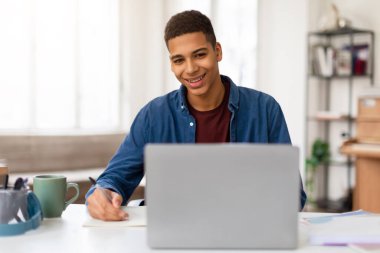 Neşeli genç adam parlak ışıklı bir odada dizüstü bilgisayarıyla çalışırken not alıyor.