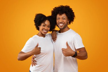 Beyaz tişörtler giyen, sarılan, baş parmağını kaldıran, kameraya gülümseyen, sarı arka planda izole edilmiş genç siyahi erkek ve kadın pozitifler. Mutlu Afrikalı Amerikalı çift güzel bir servis öneriyor.
