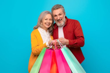 Alışveriş neşesi. Bir sürü alışveriş çantası tutan neşeli, olgun bir çift Kara Cuma anlaşmalarını ve satış tekliflerini kucaklıyor, mavi arka planda çekilmiş bir portre. Mevsimlik eğlencelerin mutluluğu