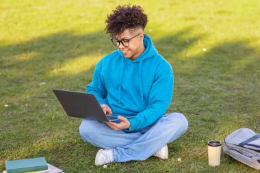 Casual young african american man in a bright blue hoodie sitting cross-legged in a grassy field, working on a laptop with a coffee cup nearby clipart