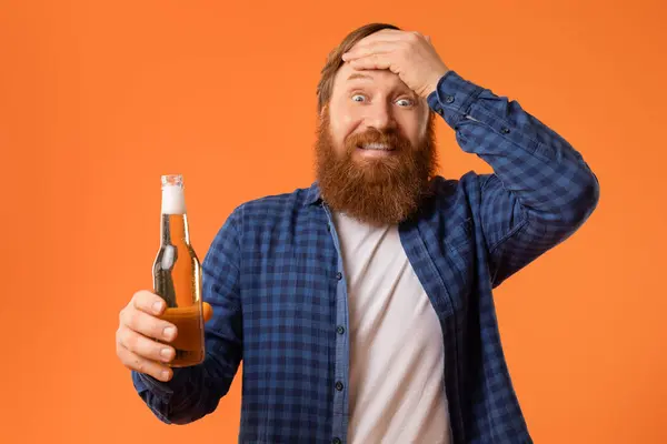 赤い毛およびひげが付いている陽気で面白い男の肖像画 ビールのびんを握り 感情的に頭に触れること スタジオのオレンジの背景 楽しみな党およびアルコール飲料の上に置きます — ストック写真