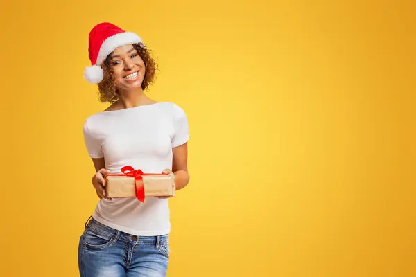 幸せなアフリカ系アメリカ人の女の子でサンタ帽子を保持クリスマスプレゼント カメラと笑顔を見て オレンジのスタジオの背景 — ストック写真