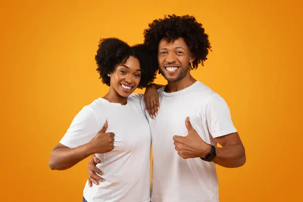 Positive young black man and woman wearing white t-shirts embracing and showing thumb up, smiling at camera, isolated on yellow studio background. Happy african american couple recommend nice service