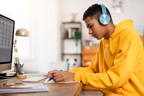 フーディーとブルーヘッドフォンに焦点を当てた10代の男はノートを書き 日当たりの良い部屋のインテリアでコンピュータで木製の机で自宅で勉強 — ストック写真