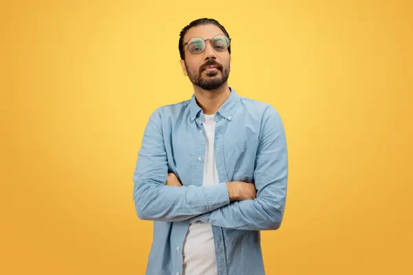一个自信的印度男人站在那里 留着胡子和眼镜 双臂交叉 穿着斜纹棉布衬衫 背景是黄色的 — 图库照片