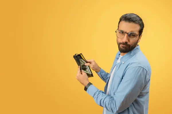 A worried indian man in a denim shirt displays an empty wallet, emphasizing financial concepts on a yellow background