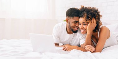 Işık dolu bir yatak odasında dizüstü bilgisayarla vakit geçiren Afro-Amerikan bir çift.