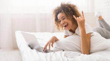 Rahat bir Afrikalı Amerikalı kadın elleriyle barış işareti gösterirken yatağının konforundan dizüstü bilgisayarı üzerinde çalışıyor.