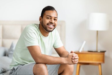 Afro-Amerikalı neşeli bir adam yatağının kenarına oturmuş elinde dijital bir tabletle sıcak, misafirperver bir gülümseme ile.