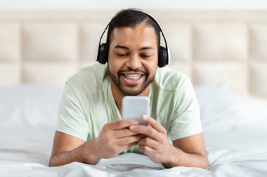 Kablosuz kulaklıklı neşeli Afro-Amerikalı adam akıllı telefon kullanıyor, eğleniyor ve rahatlıyor, yatak odasının içinde.