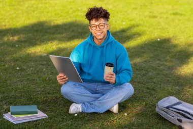 Elinde tablet ve kahve fincanı olan gülümseyen genç Brezilyalı bir adam yeşil çimlerin üzerinde kitap ve sırt çantasıyla oturuyor.