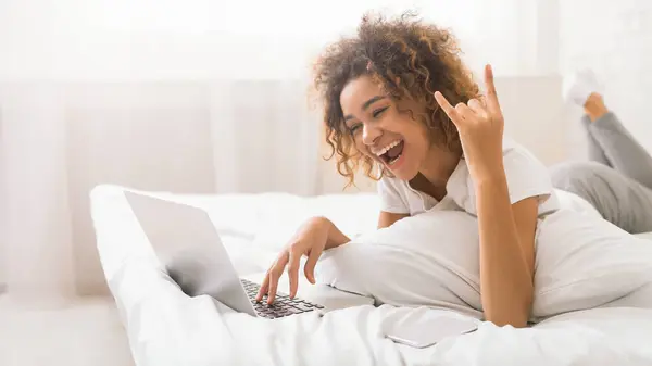 A laid-back african american woman works on her laptop from the comfort of her bed while flashing a peace sign with her hand