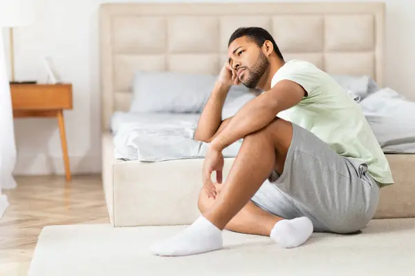 An image capturing depressed black man in casual attire sitting cross-legged on the carpet beside a bed in a cozy bedroom setting