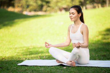 Beyazlar içindeki beyaz tenli kadın sporcu Lotus Poz verirken gözleri kapalı yoga minderinde, dışarıda güneşle ıslanmış bir parkta meditasyon yapıyor.